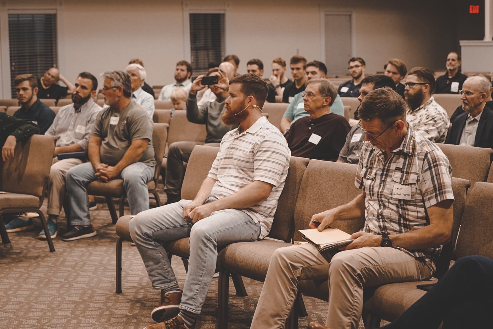 Men seated together at the Micah Mandate Weekly Rally Point, one wearing a headset microphone